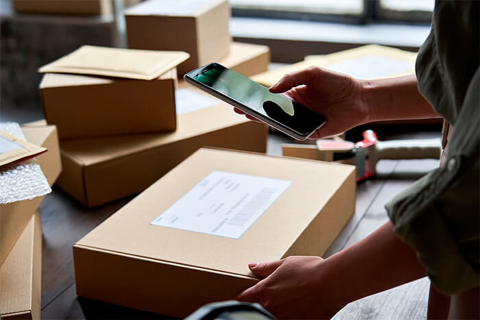 mulher com um celular na mão e uma embalagem de papelão se preparando para enviar para o produto para o cliente