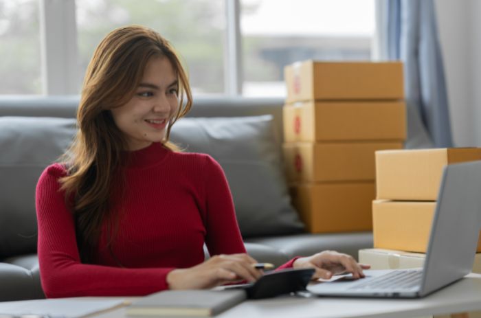 Mulher sentado à mesa, com várias caixas ao redor, notebook e calculadora, para calcular custos logísticos.